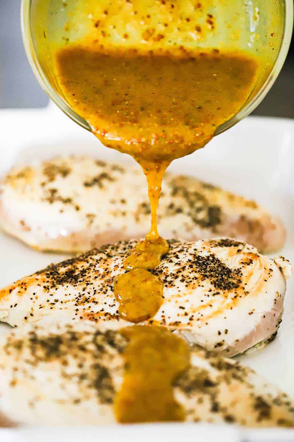 A bowl filled with honey mustard sauce being poured over seared chicken breasts in a white baking dish.