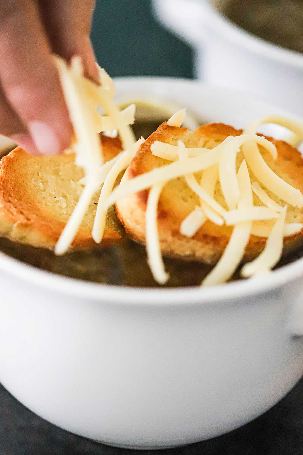 A hand sprinkling shredded Gruyere cheese over two slices of a baguette that have been toasted and sitting on a bowl of soup.