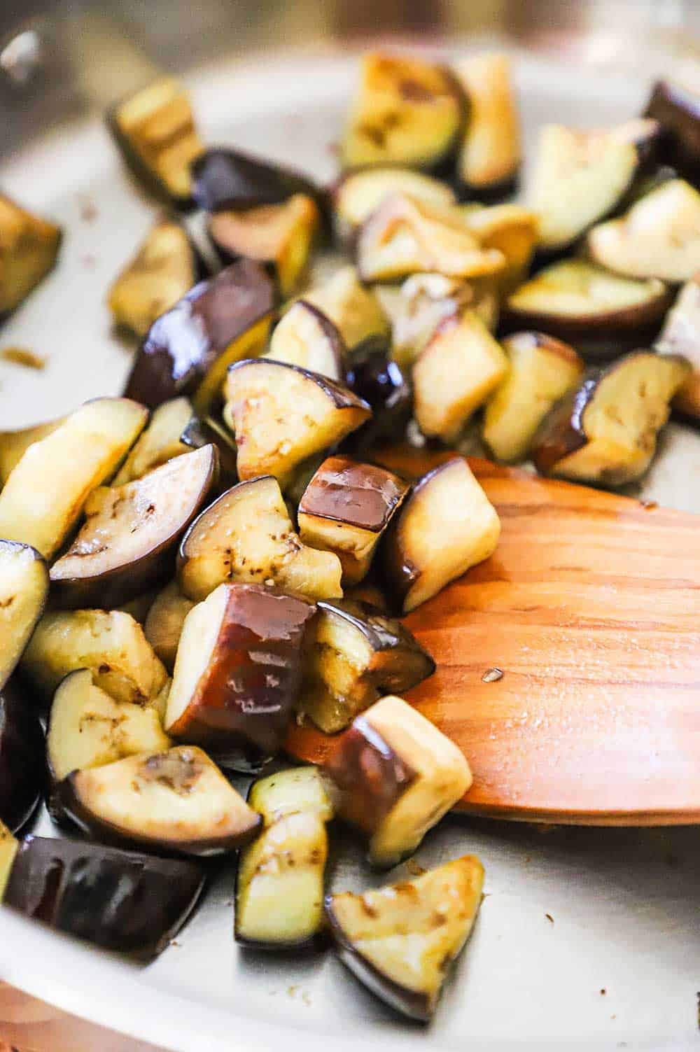 Pieces of eggplant that have been sautéed in a skillet with a wooden spatula scooting them around. 