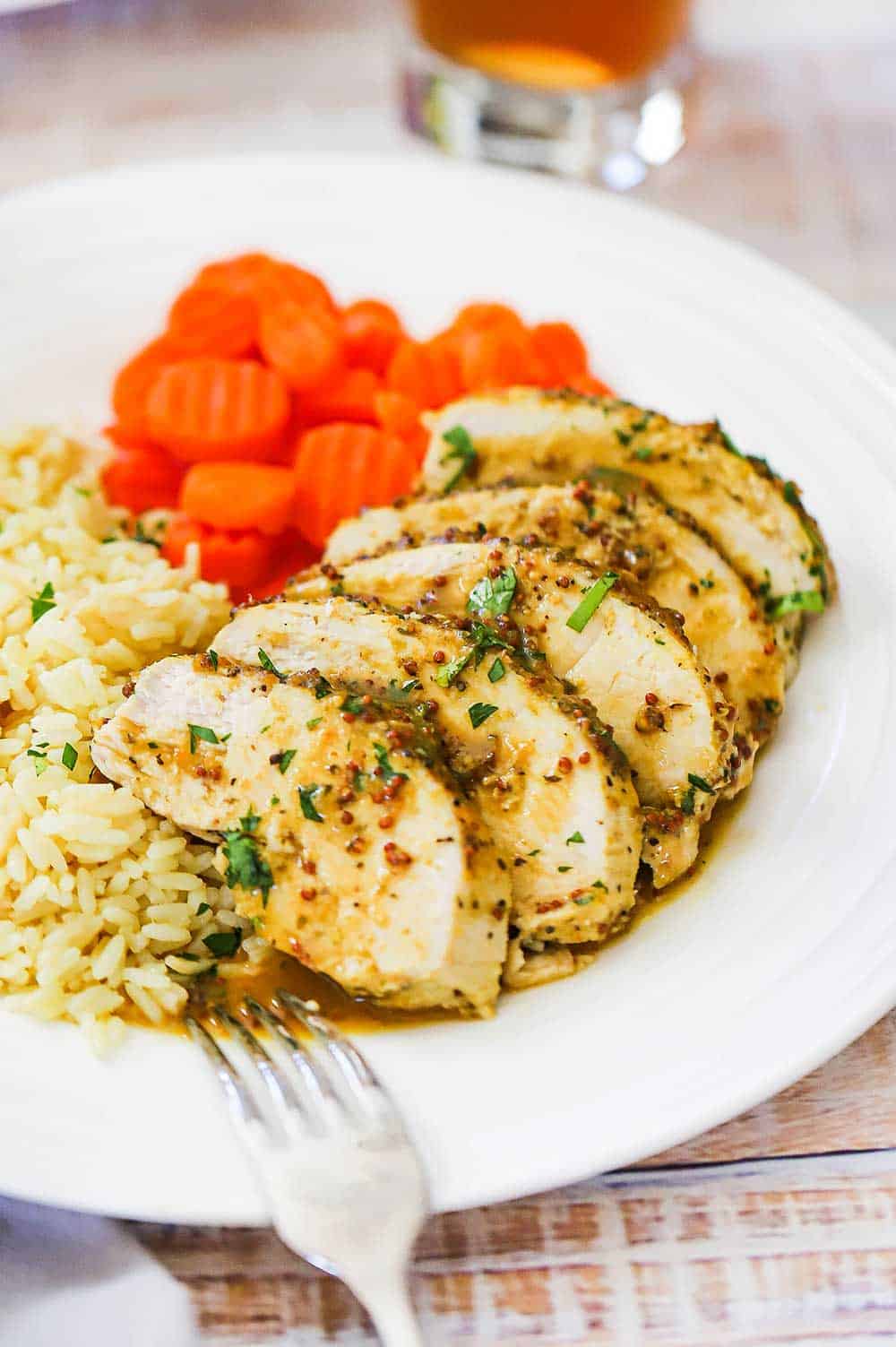A white dinner plate filled with slices of baked honey mustard chicken, sliced carrots, rice pilaf, next to a gold fork and a glass of iced tea.