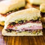 A quarter slice of a muffuletta sandwich on a cutting board surrounded by other slices and a mug of beer.