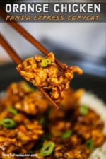 A pair of chopsticks holding up a piece of fried orange chicken over a plate of the same.