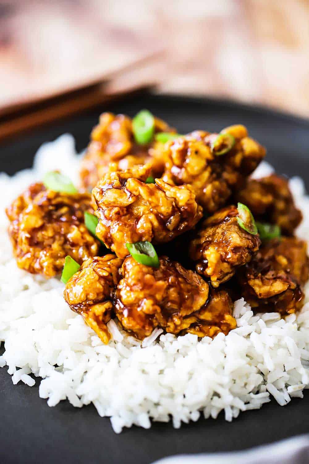 A black dinner plate filled with white rice topped with orange chicken and garnished with chopped scallions.