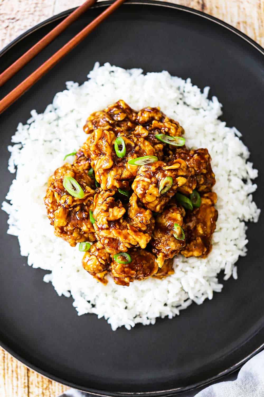 An overhead view of a black dinner plate filled with white rice topped with orange chicken with a pair of chopsticks on the side of the plate.