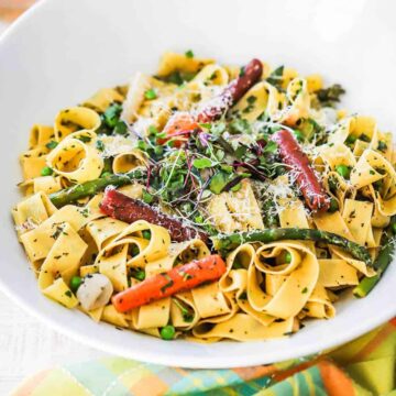A circular white serving bowl filled with pappardelle with seasonal vegetables next to a bottle of Rosé and two glasses of wine.