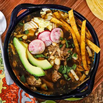 A colorful Mexican bowl filled with Mexican pozole stew surrounded by bowls of sliced radishes, cabbage, avocados, and corn tortillas.