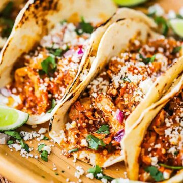 A row of easy chicken tinga tacos sitting next to each other on a wooden platter topped with crumbled cheese and garnish with lime wedges.
