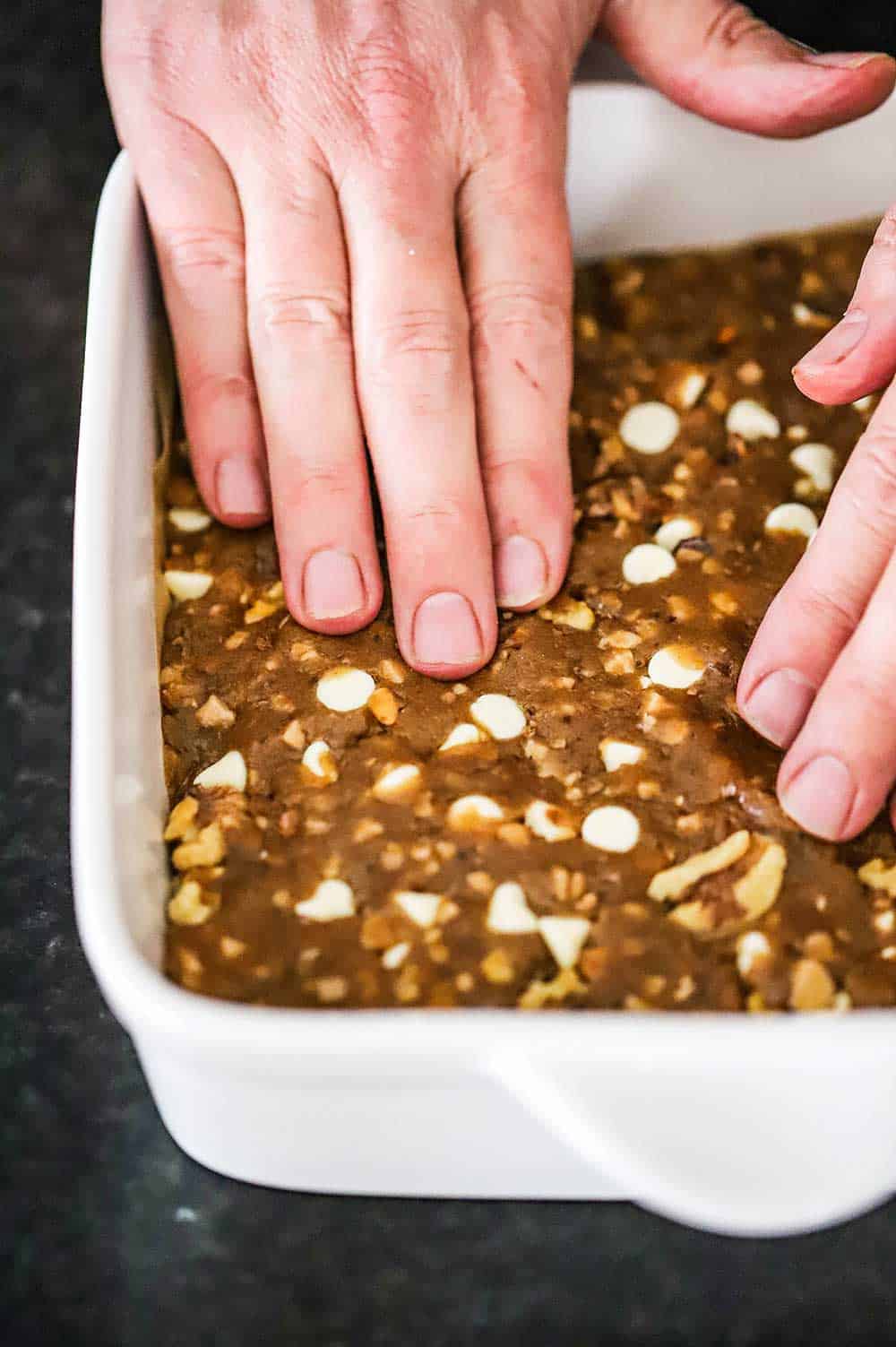 A pair of hands pressing unbaked brownie dough into a white square baking dish.