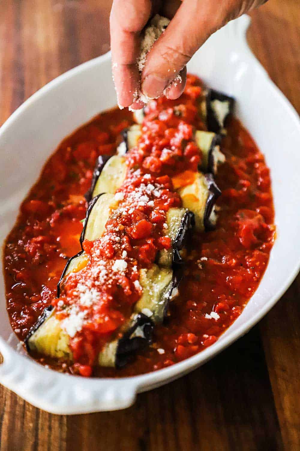 A person sprinkling grated Parmesan cheese over the top of baked eggplant rollatini in an oval baking dish.