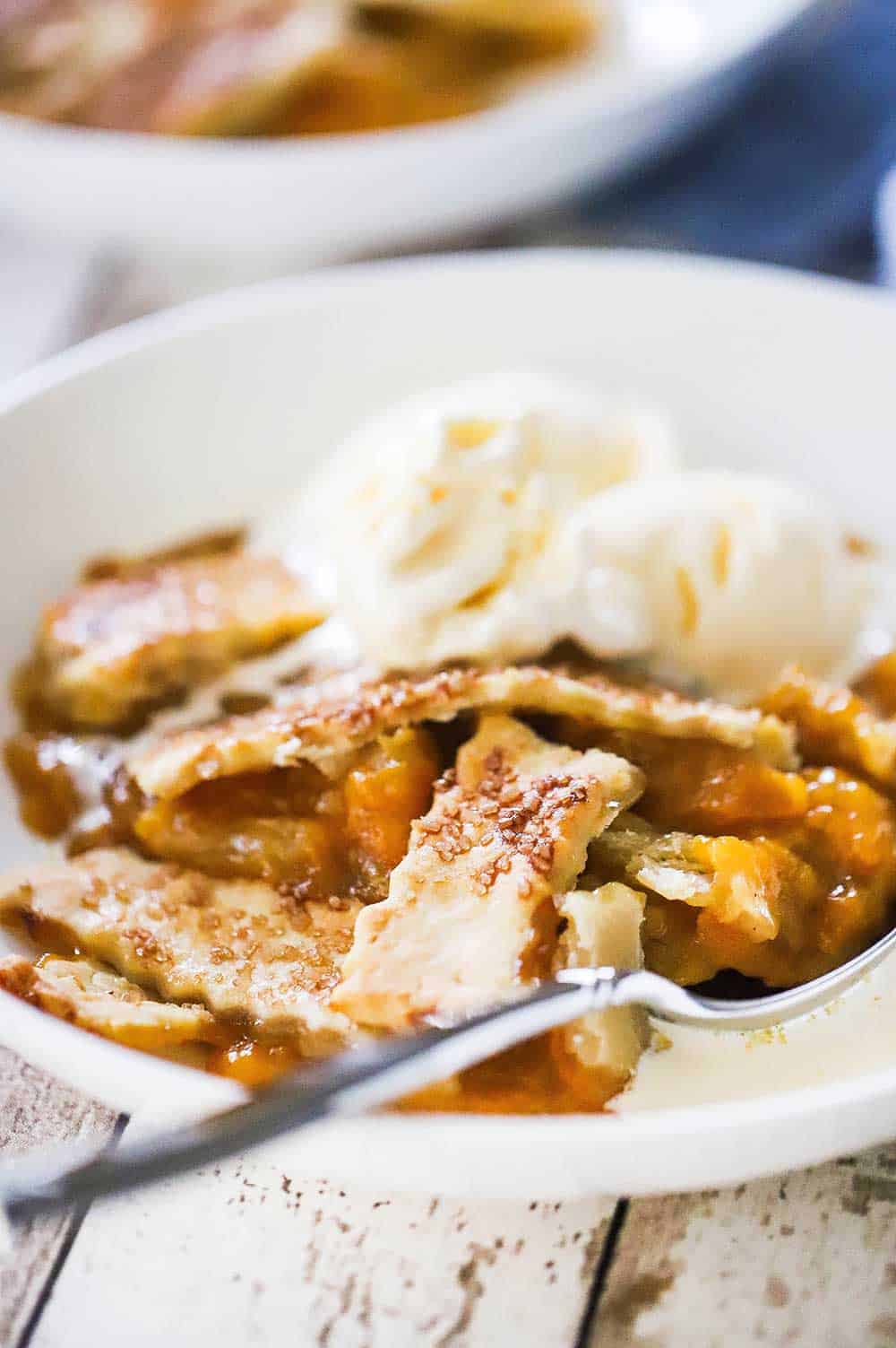 A side view of a white dessert bowl filled with old fashioned peach cobble with crust and two scoops of vanilla ice cream on top.