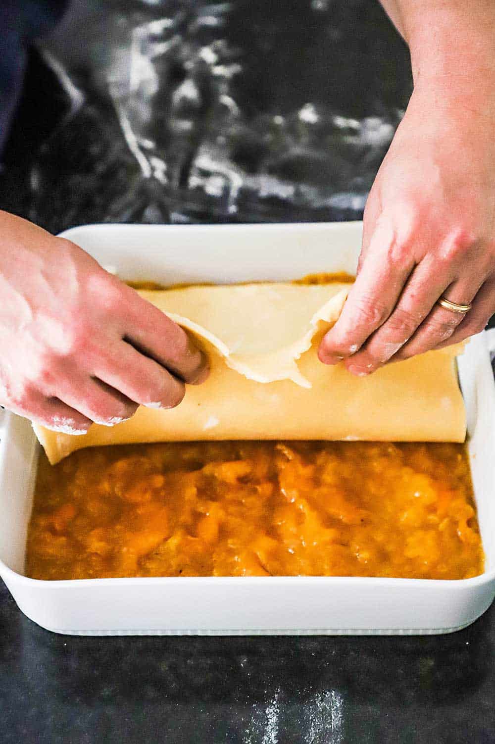 A person using their hands to layer a square piece of pie dough into a square dish that is layered with peach cobbler filling.