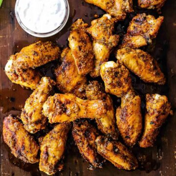 An overhead view of wings that are seasoned and grilled piled on a cutting board next to a jar of blue cheese dressing.