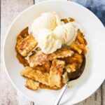 An overhead view of a white circular dessert bowl filled with old-fashioned peach cobbler and topped with three scoops of vanilla ice cream.