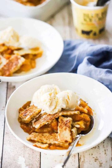 Two white bowls filled with peach cobbler and topped with scoops of vanilla ice cream with a pint of ice cream in the background.