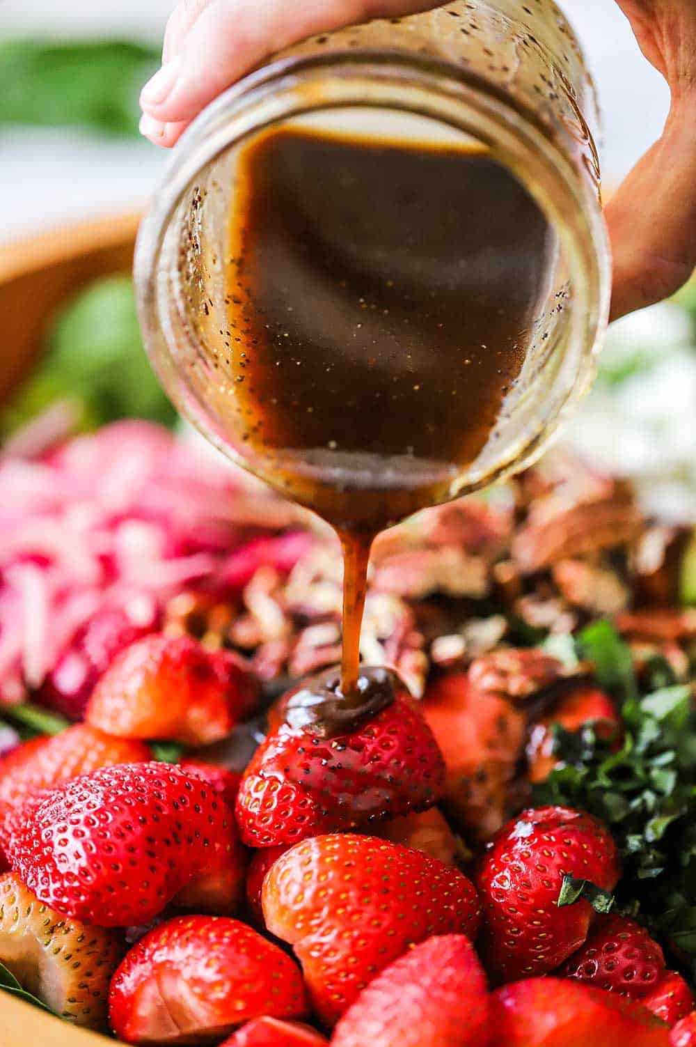 A person pouring a balsamic vinaigrette from a jar over halved strawberries in a salad bowl.