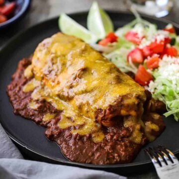 A blue plate filled with a smothered beef burrito with chili con carne sitting next to a salad topped with diced tomatoes.