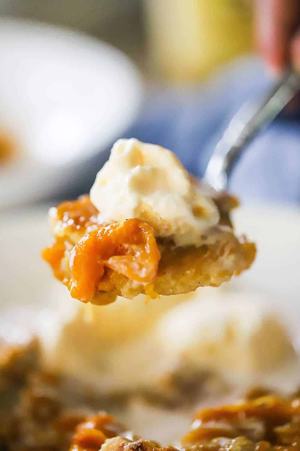 A person holding up a large spoonful of baked peach cobbler topped with vanilla ice cream.