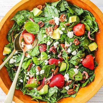 An overhead view of a fresh strawberry spinach salad with avocado that has been tossed together in a wooden bowl.