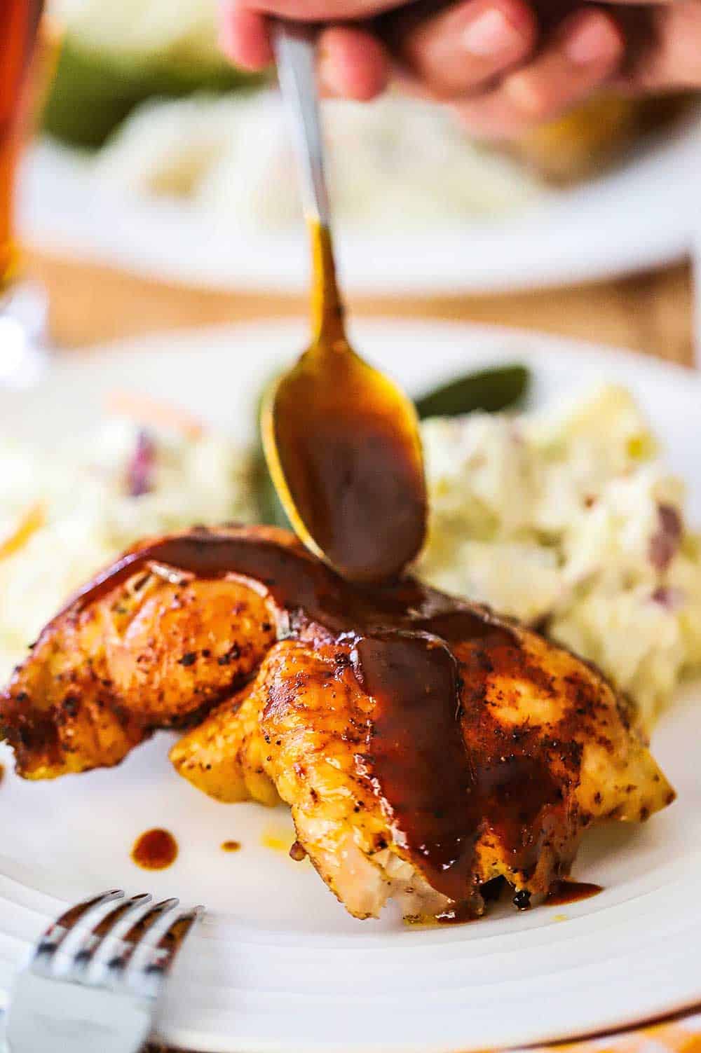 A person using a spoon to drizzle barbecue sauce over a grilled beer can chicken leg quarter.
