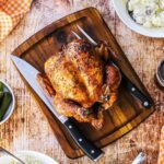 A grilled whole beer-can chicken sitting on a cutting board next to a carving knife and a bowl of BBQ sauce