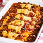 A chili cheese dog casserole topped with crumbled bacon in a white baking dish.