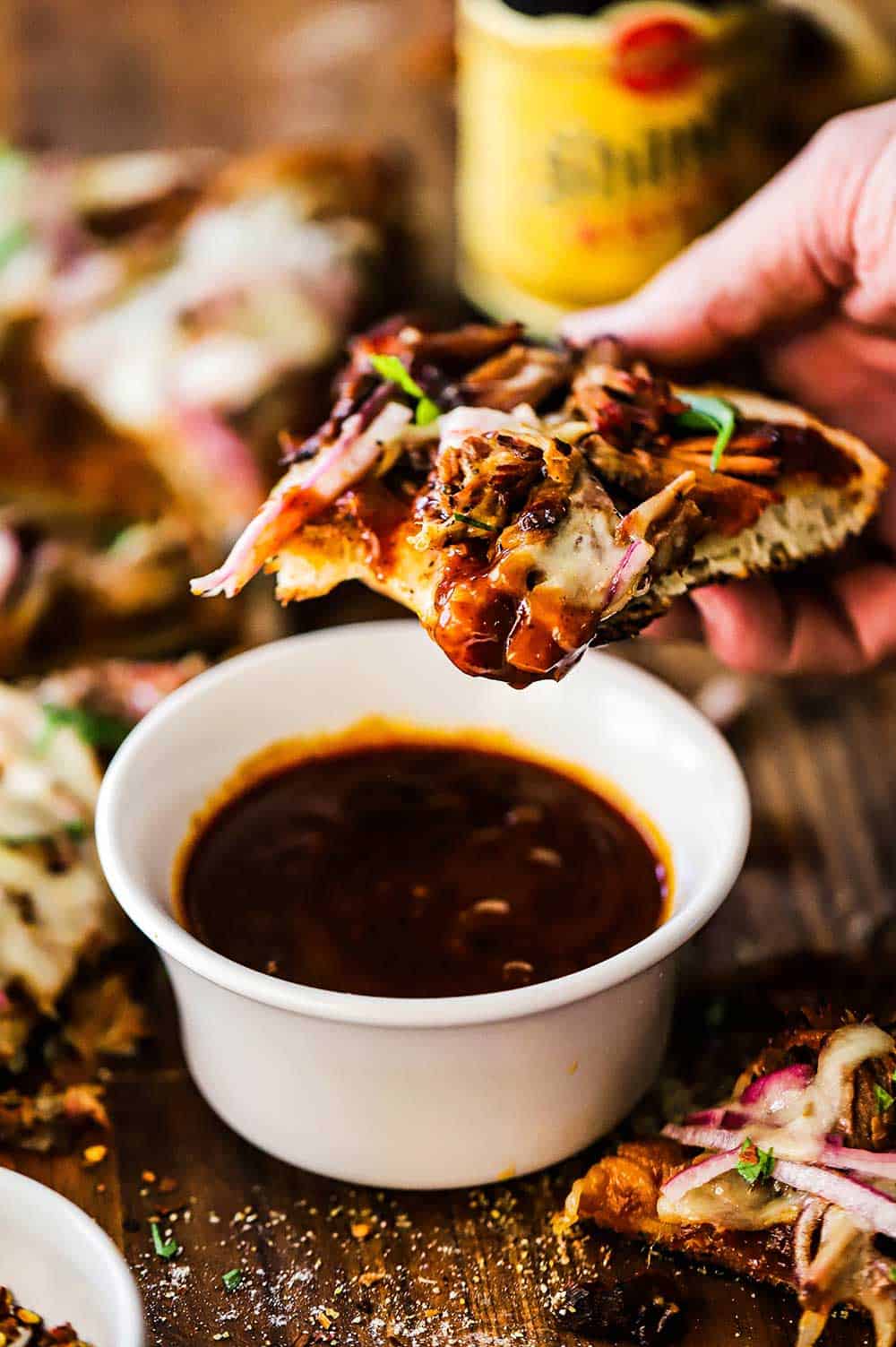 A person holding a slice of pulled pork pizza with a bite taken out of it and held above a small bowl of barbecue sauce.