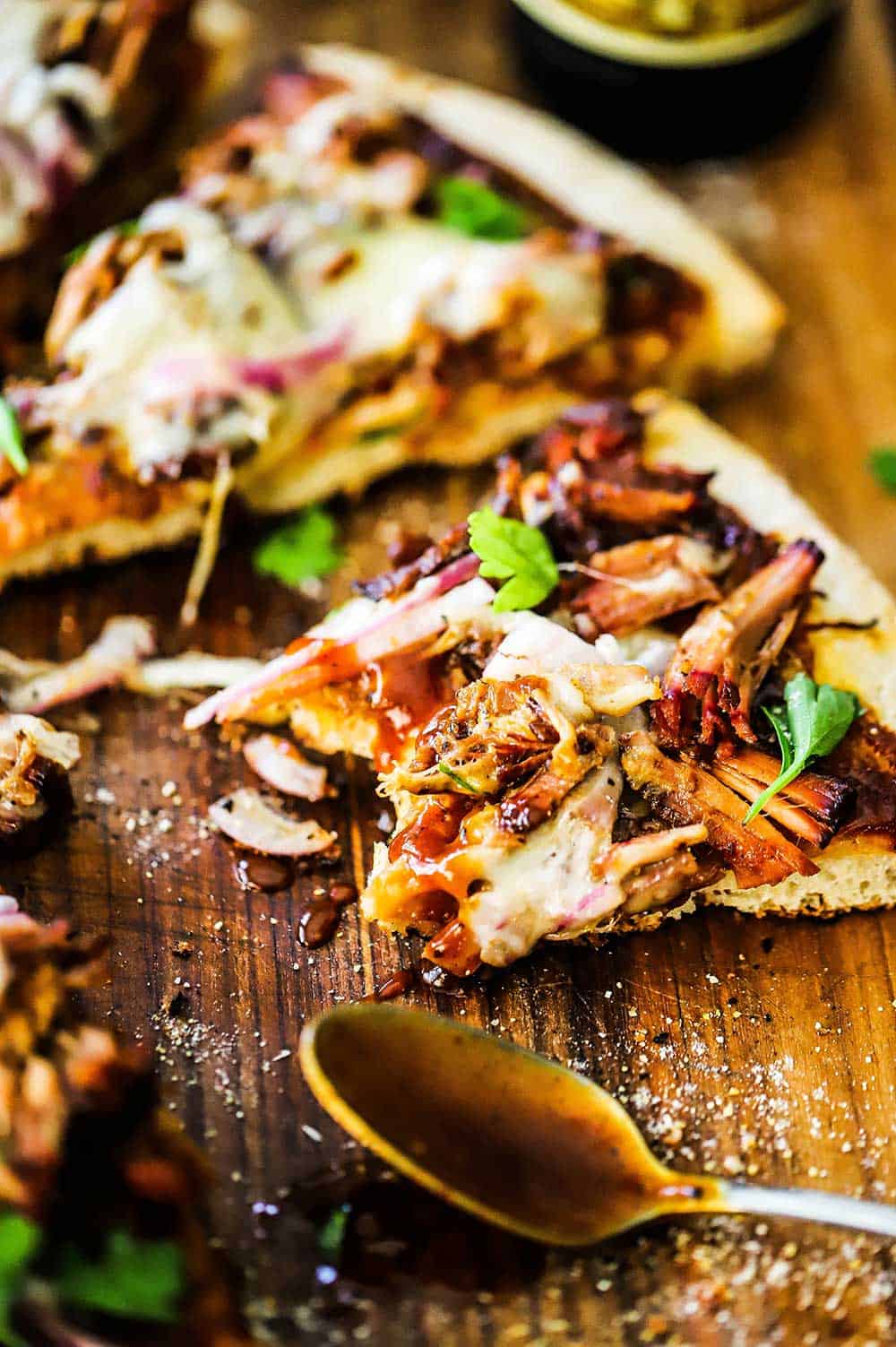 A slice of pulled pork pizza with a bite taken out of it sitting on a cutting board next to a spoon covered in barbecue sauce.