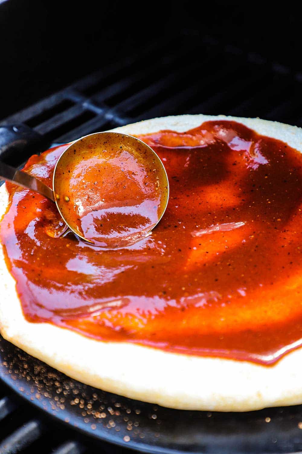 A person using a small ladle to spread barbecue sauce all over a pizza dough that is cooking on a stone on a grill.
