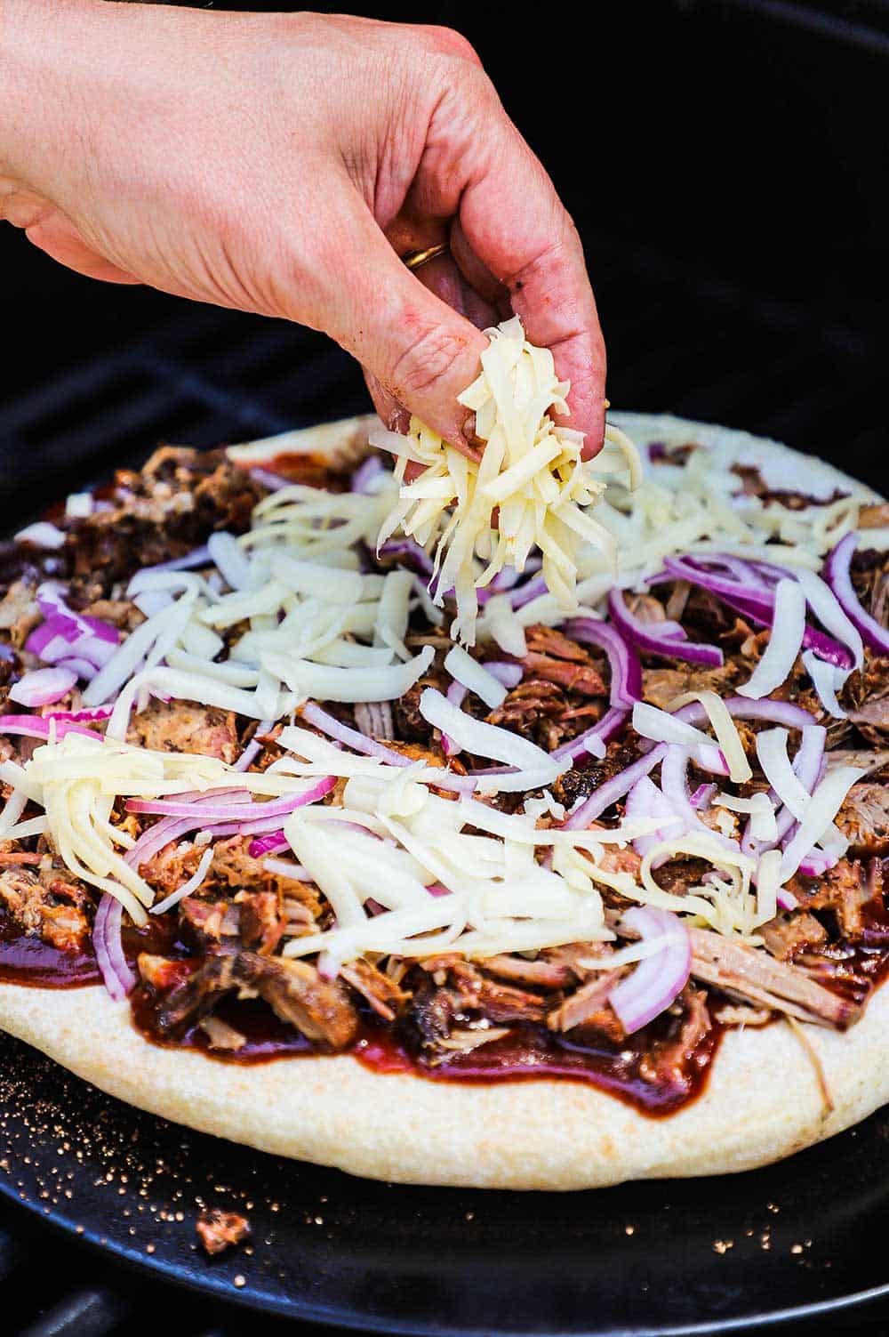 A person sprinkling shredded white cheese over a pizza dough covered with barbecue sauce, pulled pork, red onion.