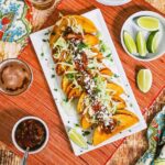 An overhead view of a white rectangular platter filled with a lineup of shrimp tacos dorados and surrounded by limes, salsa, and a glass of beer.