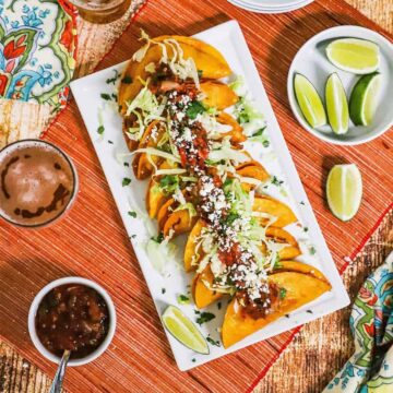 An overhead view of a white rectangular platter filled with a lineup of shrimp tacos dorados and surrounded by limes, salsa, and a glass of beer.