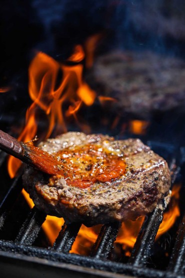 A person brushing on Korean BBQ sauce onto a beef hamburger patty that is being flame-grilled on a gas grill. 