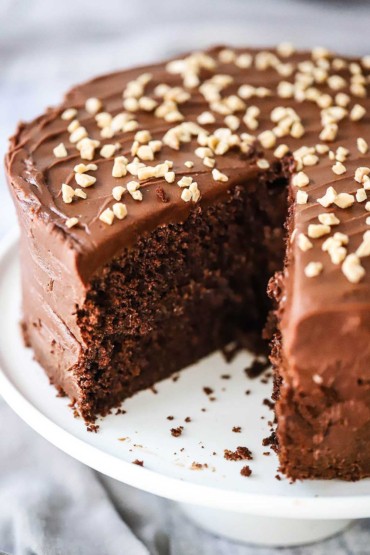 A classic chocolate cake sitting on a white cake stand and is topped with toffee bits and a slice of the cake has been removed.