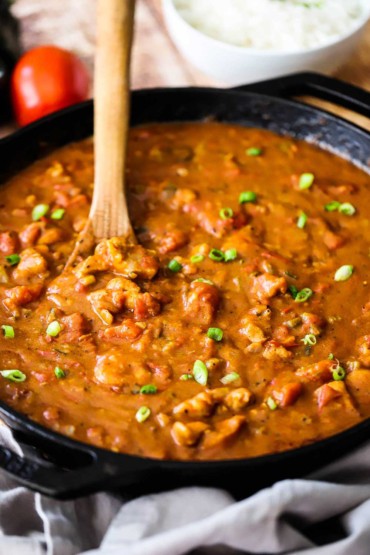 A large black cast-iron skillet filled with a chicken creole sauce with a wooden spoon inserted into the middle.