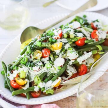 A straight-on view of a green bean and tomato salad with scallion dressing on an oval white platter next to a glass of white wine.