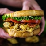 A person holding a Fried Oyster BLT consisting of two toasted slices of bread with crunchy oysters, crispy bacon, sliced tomatoes, and lettuce.