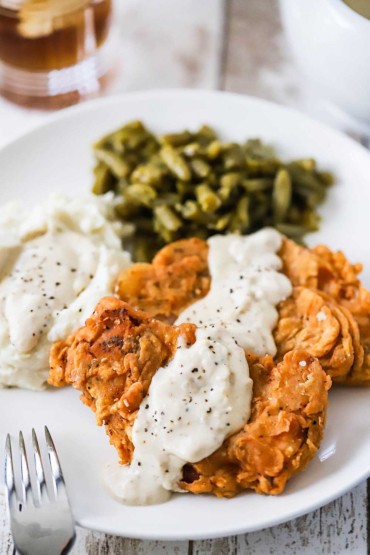 A close-up view of two Southern-fried pork chops on a dinner plate with cream gravy drizzled over the top of them and sitting next to mashed potatoes and green beans.