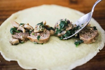 A spoon being used to transferred cooked pieces of Italian sausage and sautéed spinach and mushrooms onto a crêpe.