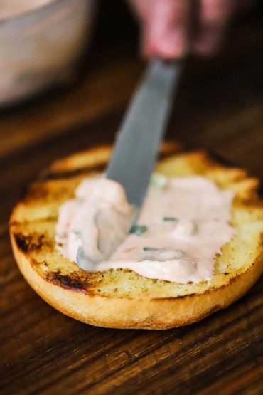 A person using a knife to spread chili sauce mayonnaise onto a toasted brioche bun.