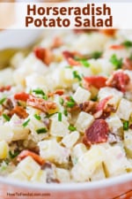 A close-up view of a white bowl filled with creamy horseradish potato salad topped with bacon pieces and snipped chives.