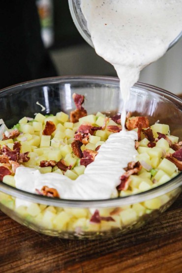 A large glass bowl filled with cooked cubed potatoes and pieces of bacon with white horseradish dressing being poured over the top.
