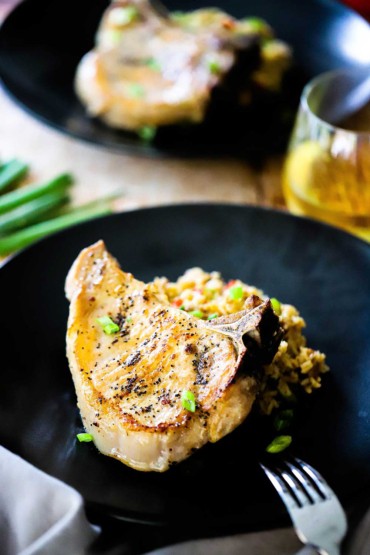 A pan-seared pork chop sitting on a bed of rice on a black dinner plate with a glass of white wine nearby.