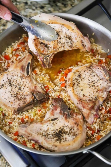 A person using a pair of metal tongs to place a partially cooked bone-in pork chop into a skillet filled with other chops and rice and veggies in a broth.