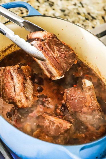 A person using a pair of tongs to place a beef short rib into a large Dutch oven filled with other short ribs and a tomato and vegetable sauce. 