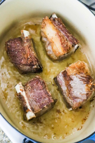 Four beef short ribs that are being seared in an oval Dutch oven with olive oil. 