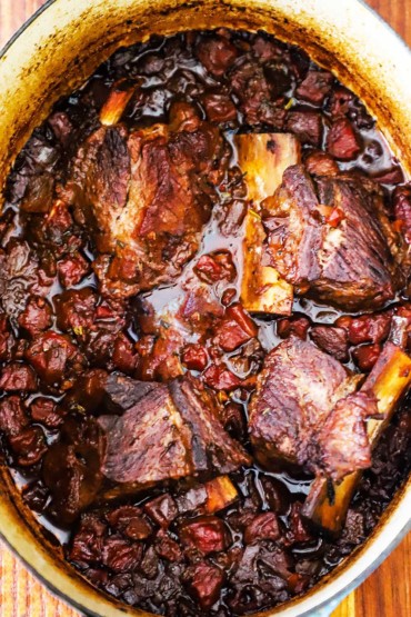 An overhead view of a large oval Dutch oven that is filled with braised beef short ribs in an Italian tomato and vegetable sauce. 