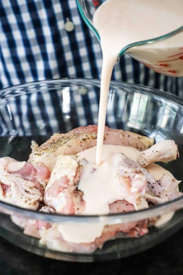 A person pouring buttermilk from a large glass measuring cup into a glass bowl filled with seasoned uncooked chicken pieces.