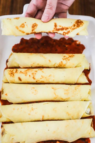 A person using his hand to place a cheese-stuffed crêpe in a casserole dish lined with marinara sauce.