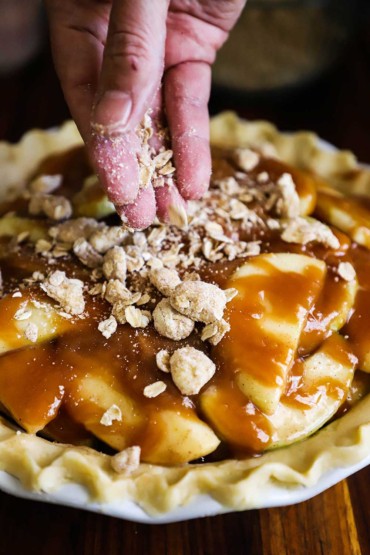 A hand sprinkling an oats, spice, and butter crumble over the top of an unbaked caramel apple pie. 