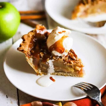 A slice of caramel apple pie on a white dessert plate topped with a scoop of vanilla ice cream and a drizzle of caramel sauce.